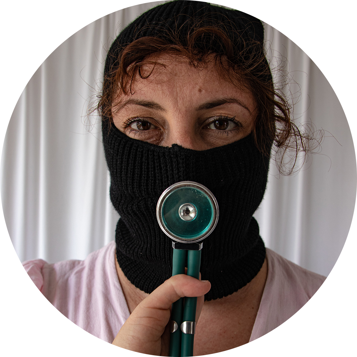 Fotografia colorida de uma mulher branca com um capuz preto sobre o rosto. Somente os olhos castanhos e parte da testa estão à mostra. Na altura da boca, sobreposto ao capuz, está pendurada a parte circular de um estetoscópio, do qual pendem dois tubos plásticos. O equipamento é azul-claro, com detalhes cinza. 
