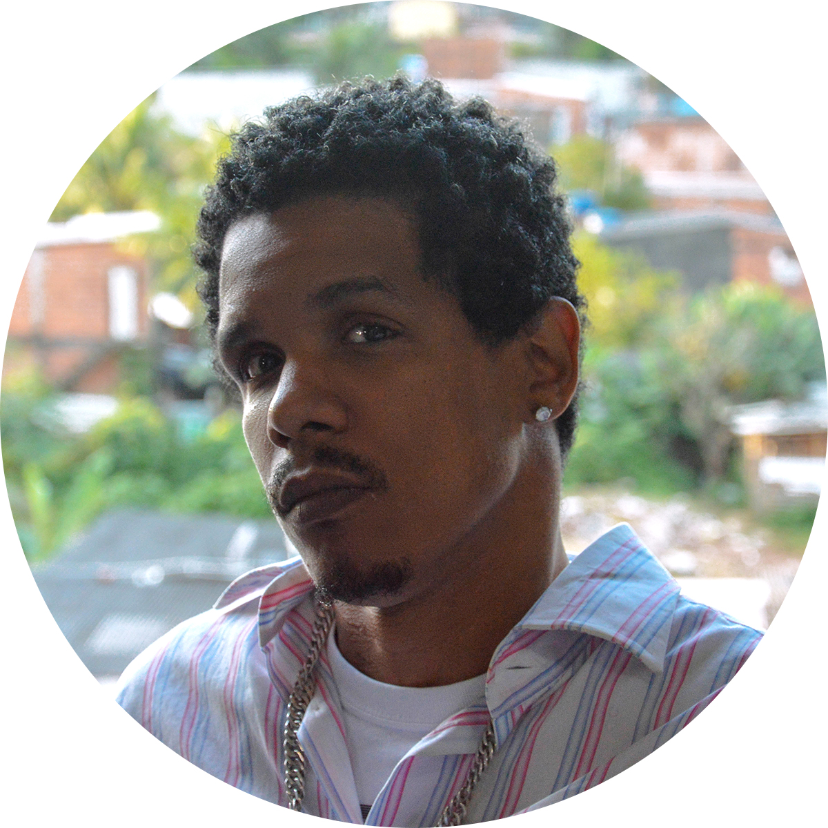 Fotografia colorida de MC Deisdetih, um homem negro, de cabelo preto, crespo e curto, olhos castanhos, nariz largo, lábios grossos e bigode e barbicha curtos. Ele aparece de perfil, voltado para a esquerda da foto, olhando de canto para a nossa direção com uma expressão séria. Usa um brinco de brilhante na orelha esquerda, uma corrente longa e de prata por cima de uma camisa branca de listras verticais vermelhas e azuis e uma camiseta branca por baixo. 
