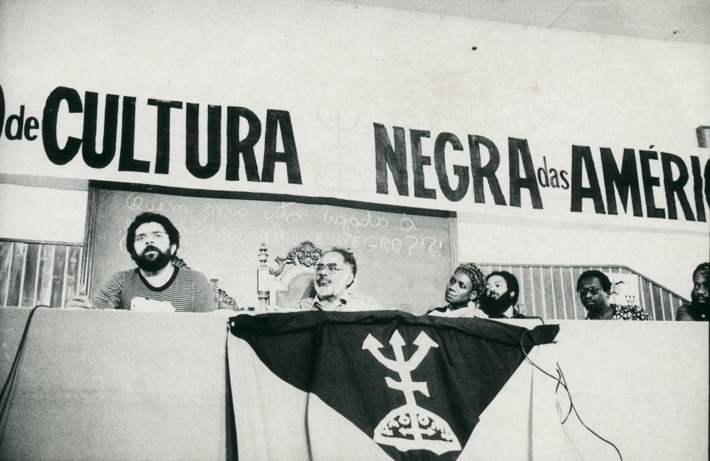 Luís Inácio Lula da Silva e Abdias Nascimento na Pontifícia Universidade Católica (PUC) de São Paulo, durante o 3º Congresso de Cultura Negra das Américas, em 1982. Na mesa, estão a ativista Esmeralda Brown, do Panamá, o líder sindical Marvin Wright Lindo, da Costa Rica, e o professor Anani Dzidzienyo, de Gana