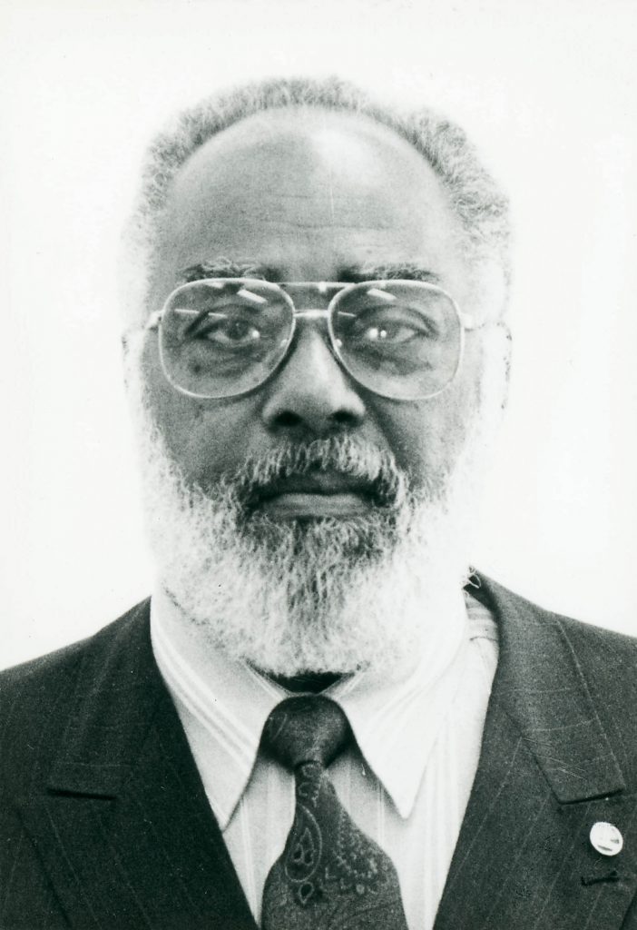 Homem negro com cabelos e barba grisalhos em foto preto e branca de documento. Ele está sério, usa óculos de grau e terno escuro, com camisa branca e gravata.