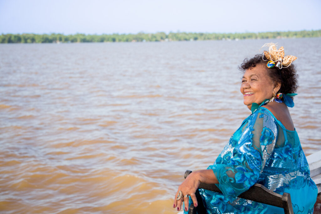 Imagem colorida de Dona Onete durante ensaio fotográfico para o álbum Banzeiro. Dona Onete está sentada, com um vestido azul. Ao fundo, o rio da cidade de Belém.